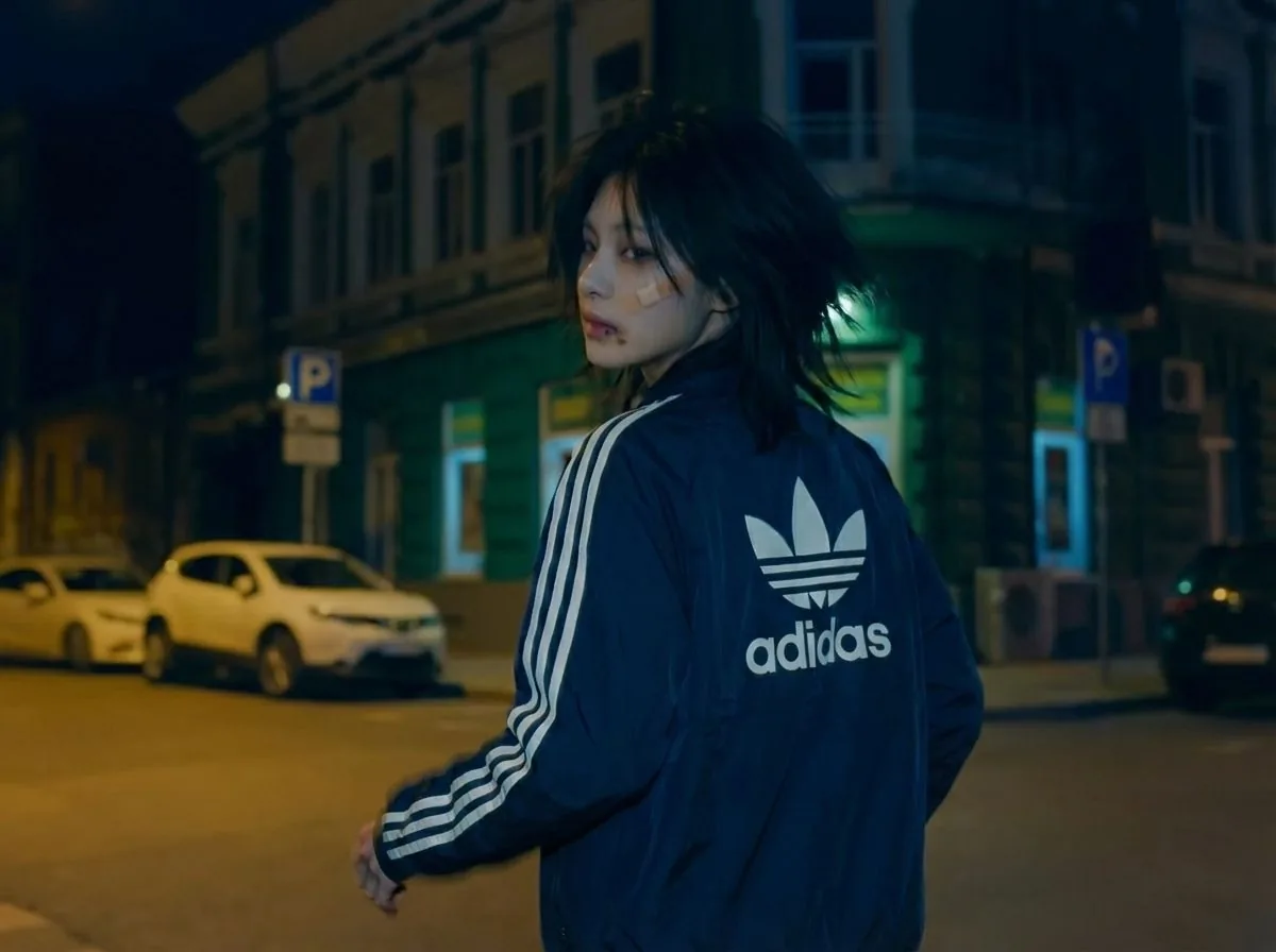 The same figure turning back toward the camera on the same street, a plaster on one cheek, the three stripes running down the sleeve.