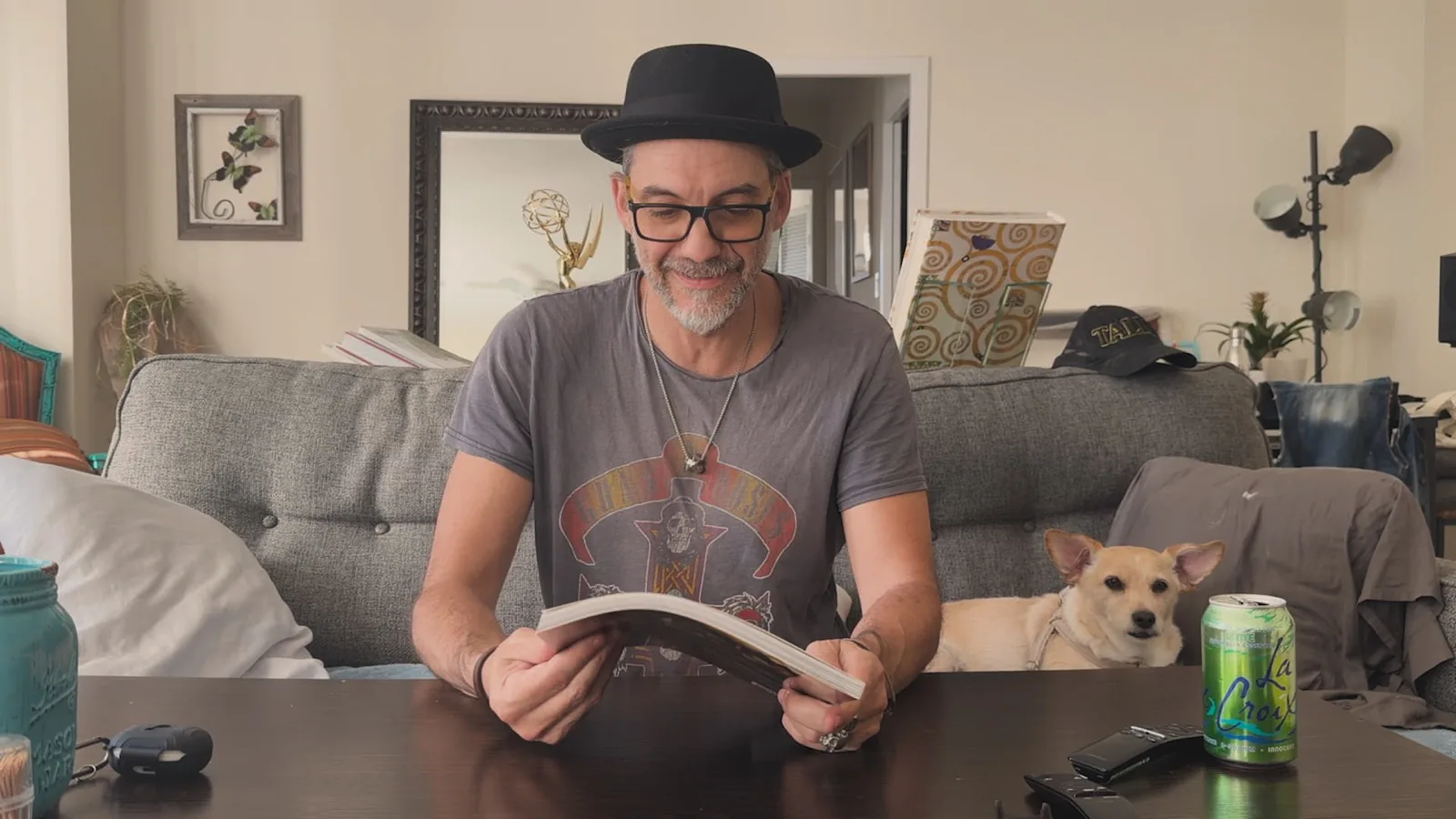 Alessandro Taini sits at his dining table in a hat and glasses, turning the pages of his own artbook, with his Emmy statuette on the shelf behind him and his dog on the couch.